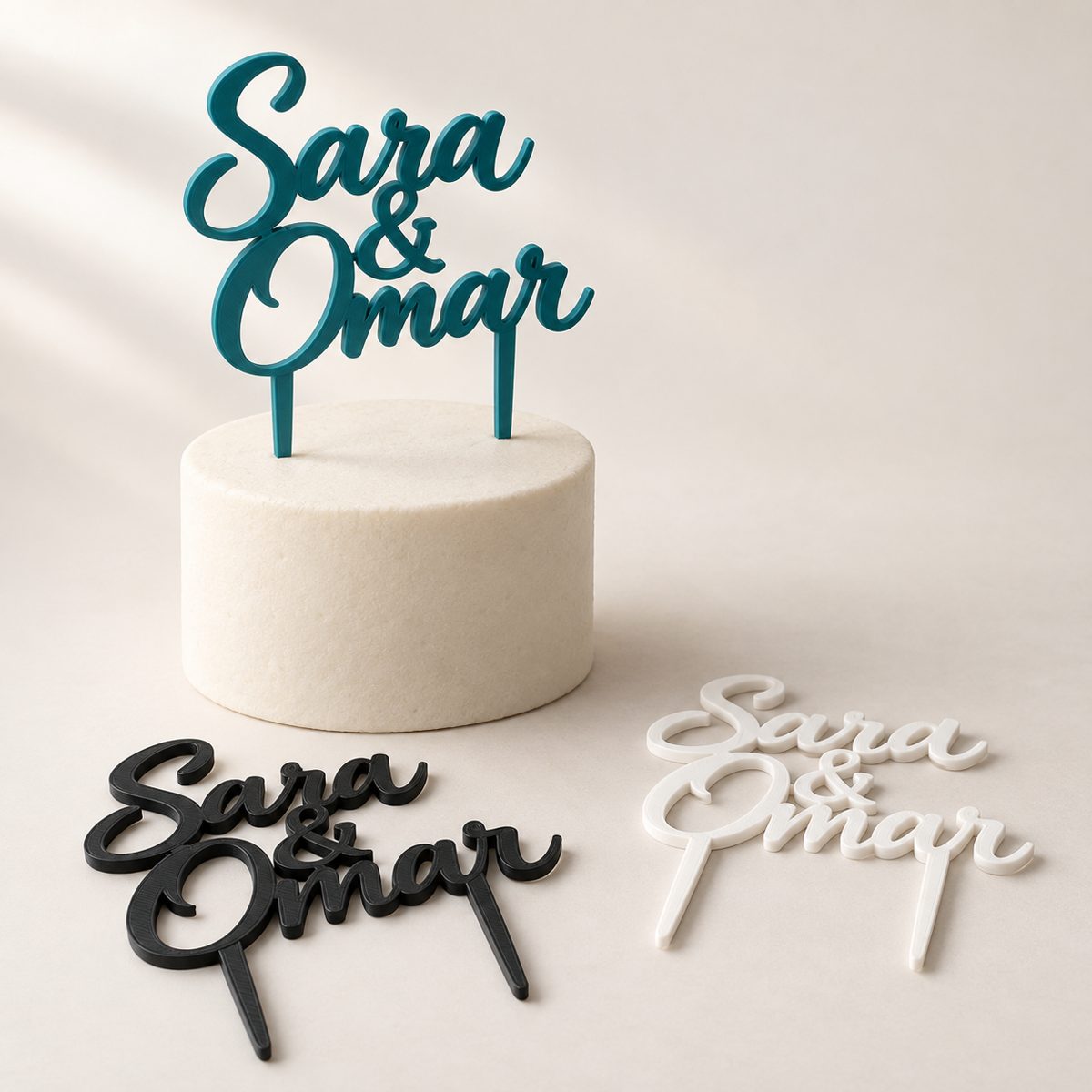 Script cake topper reading Sara & Omar in teal on a small white cake, with black and white versions beside it