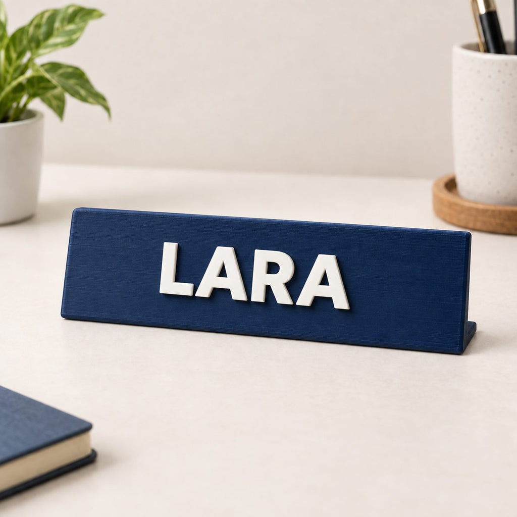 Navy desk nameplate with raised white letters spelling LARA, on a desk beside a book and a small plant