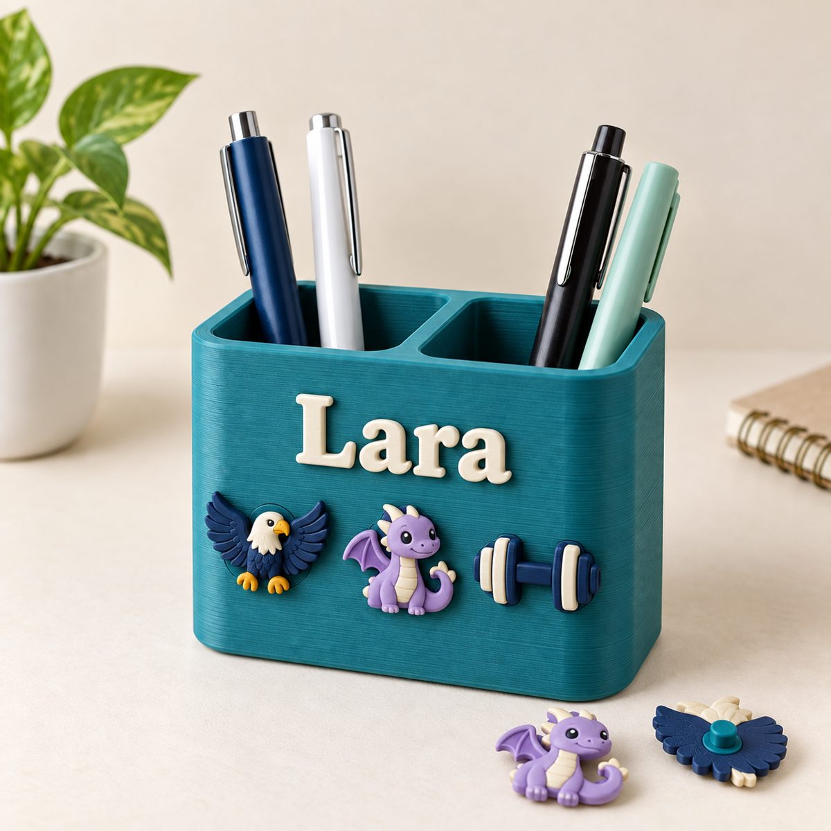 Teal two-slot pen holder with the name Lara and eagle, dragon and dumbbell charms on the front, holding pens